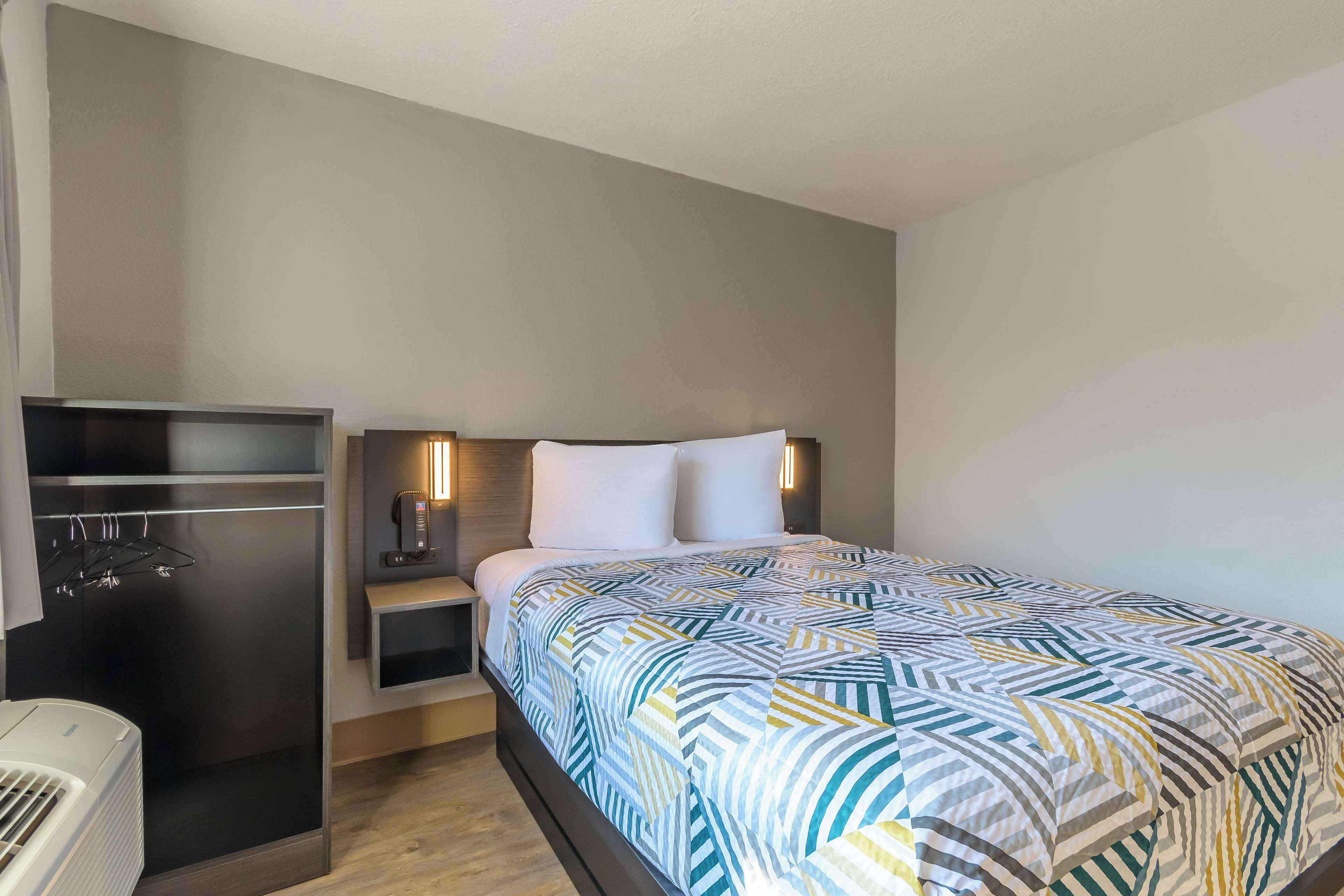 A hotel room with a bed, bedside table, and a built-in air conditioning unit.