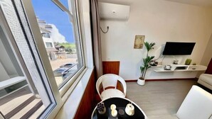 Una sala de estar moderna con un ventanal grande, un televisor de pantalla plana montado en la pared, un sofá blanco, una silla de mimbre y una pequeña mesa redonda con objetos decorativos.
