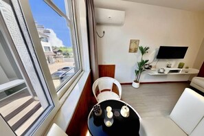 Un salon moderne doté d’une grande fenêtre, d’un téléviseur à écran plat fixé au mur, d’un canapé blanc, d’un fauteuil en osier et d’une petite table ronde agrémentée d’objets décoratifs.