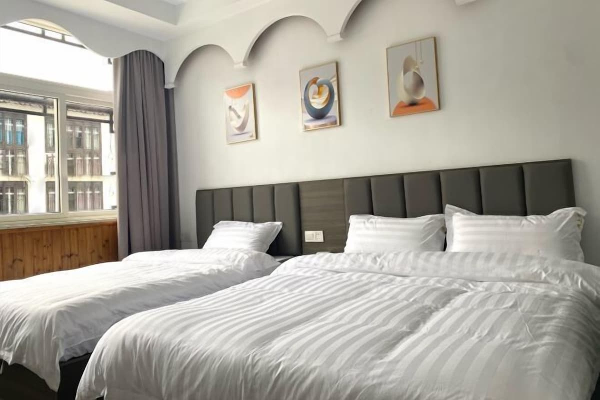 A hotel room with two beds, a large window with curtains, and three abstract paintings on the wall.