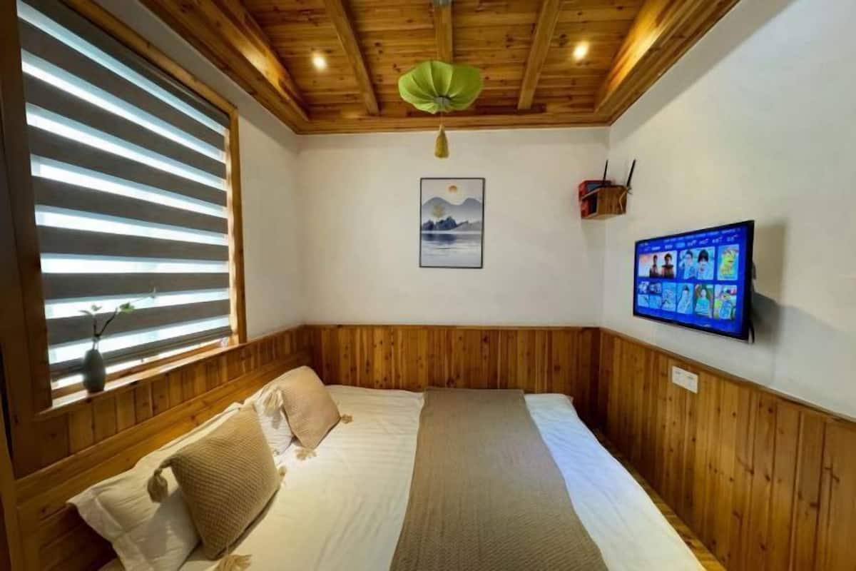A room with wooden paneling, a bed with a beige blanket, a wall-mounted TV, and a painting of mountains.