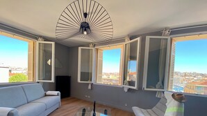 Een moderne woonkamer met een bank, een glazen salontafel en een fauteuil. Grote ramen met open luiken bieden uitzicht op de skyline.
