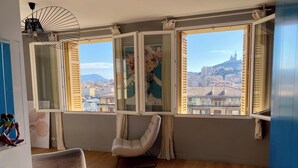 Een kamer met houten vloeren, een witte stoel en grote ramen met uitzicht op een stadsbeeld.