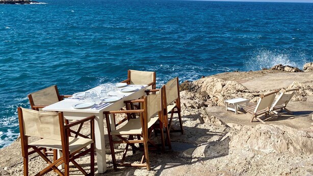 Een tafel met stoelen op een rotsachtig strand, met uitzicht op de zee en verre kliffen.