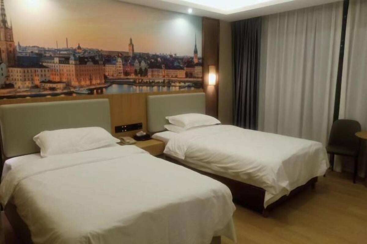 Ein Hotelzimmer mit zwei Betten, einem Nachttisch, einer Lampe, einem Sessel und einem Wandgemälde mit Stadtansicht.