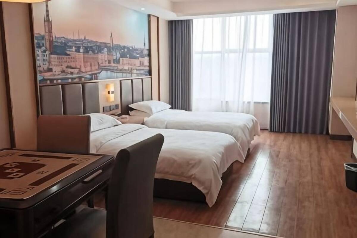 Ein Hotelzimmer mit zwei Betten, einem Schreibtisch, einem Sessel, einem Stadtbild und einem großen Fenster mit Vorhängen.