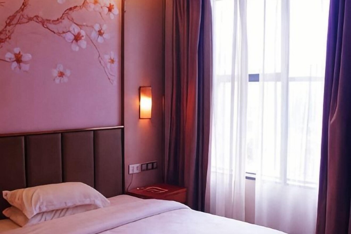 Ein Hotelzimmer mit Bett, Nachttisch, Wandlampe und einem Fenster mit Vorhängen.
