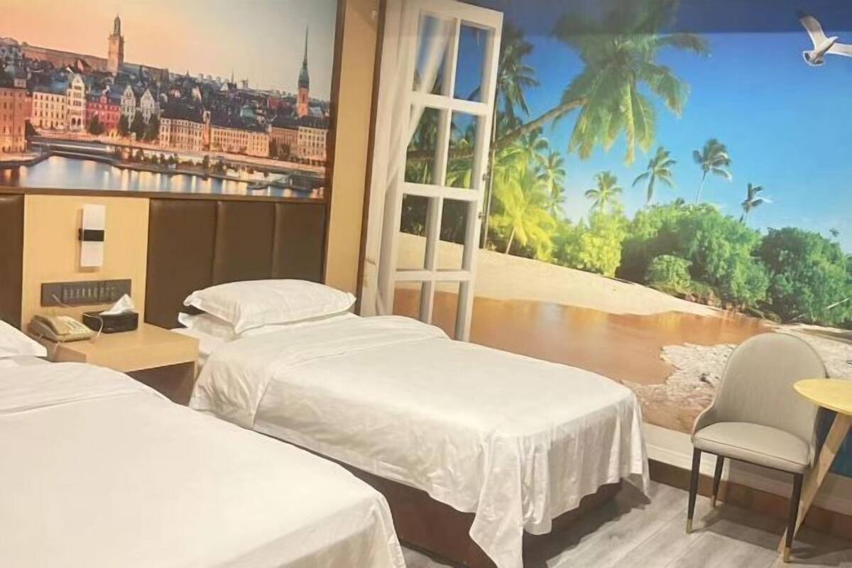 Ein Hotelzimmer mit zwei Betten, einem kleinen Tisch, einem Sessel und einem Wandgemälde von Stadt und Strand.