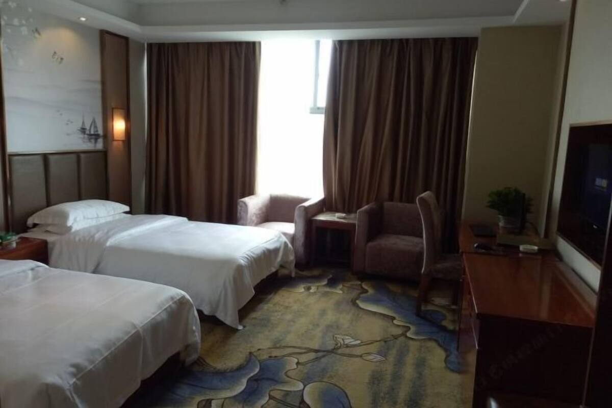 Ein Hotelzimmer mit zwei Betten, einem Fernseher, einem Schreibtisch, einem Stuhl und einem Fenster mit Vorhängen.