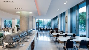 Une cafétéria moderne avec de longues tables et des chaises, un pilier central et de grandes fenêtres donnant sur un espace verdoyant.