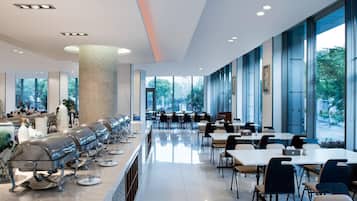 A modern cafeteria with long tables and chairs, a central pillar, and large windows offering a view of greenery.