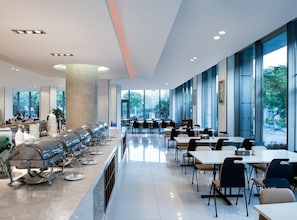 Una cafetería moderna con mesas y sillas largas, un pilar central y grandes ventanales que ofrecen vistas a zonas verdes.