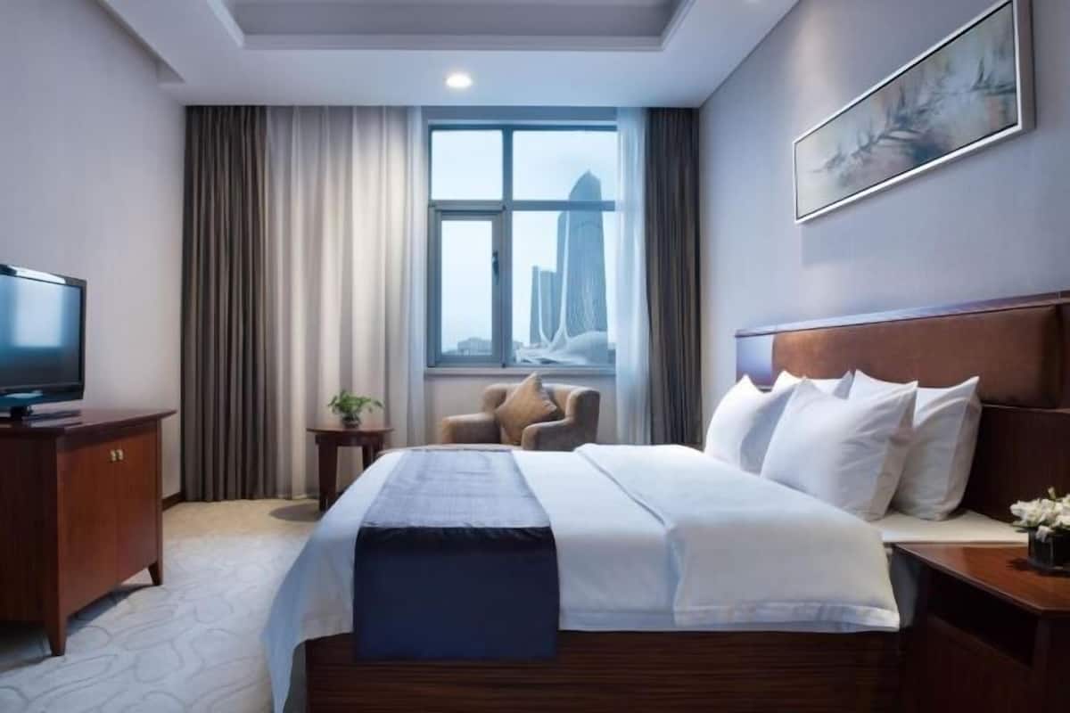 Une chambre d’hôtel avec un lit, une télévision, une fenêtre avec des rideaux, une chaise et un tableau au mur.