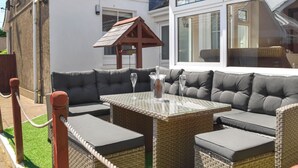 Un patio con una mesa de vidrio, asientos con cojines grises y una pequeña estructura de madera.