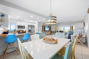 A modern kitchen with white cabinets, a dining area with a white table, blue chairs, and a hanging light fixture. - Jamaica Royale Beach View Tower by Beachside Management (Siesta Key)