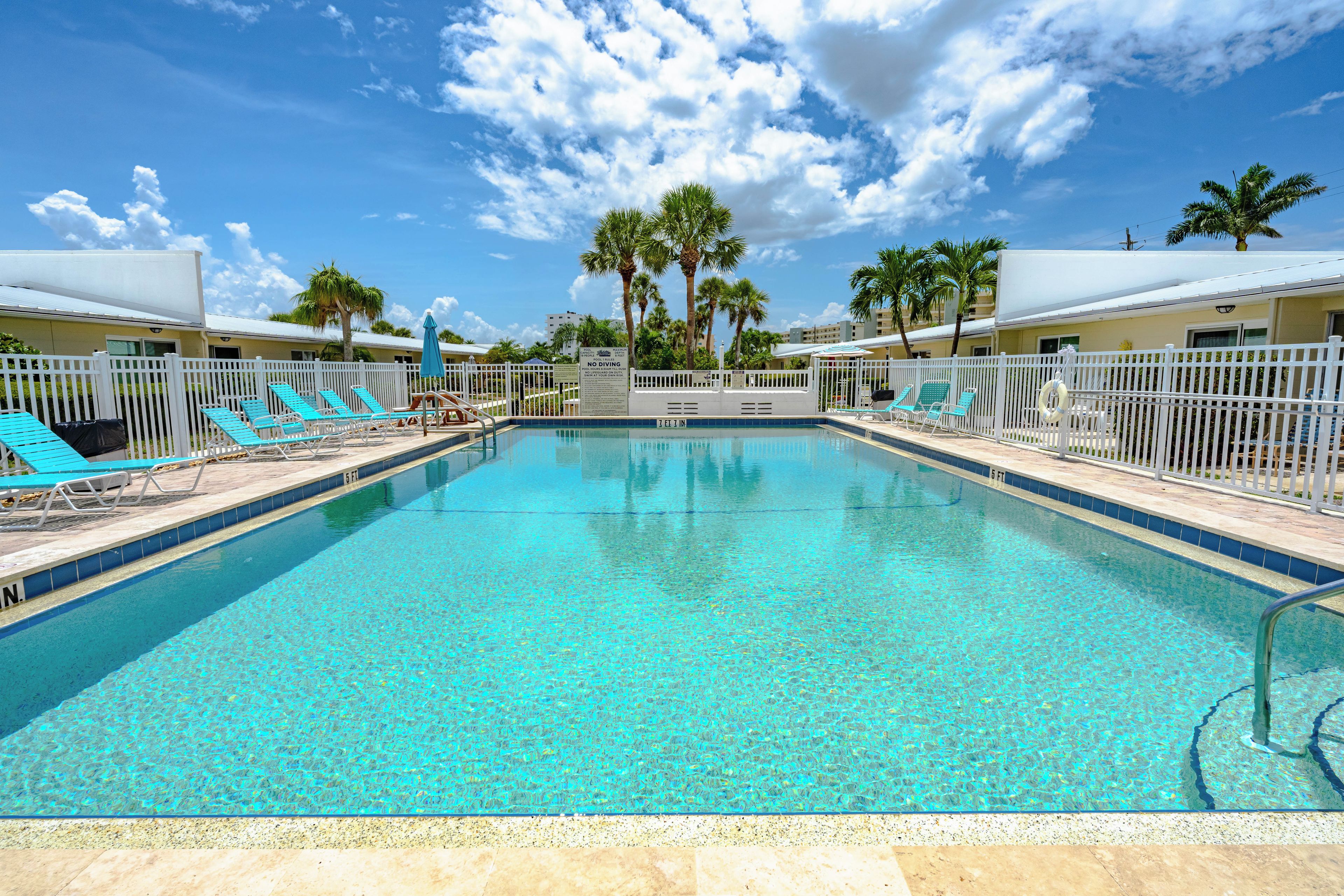 Une piscine d’un bleu éclatant, bordée de chaises longues et entourée de palmiers, le tout délimité par une clôture blanche.