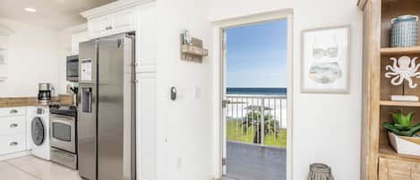A modern kitchen with stainless steel appliances, a washing machine, and a view of the sea through an open door.