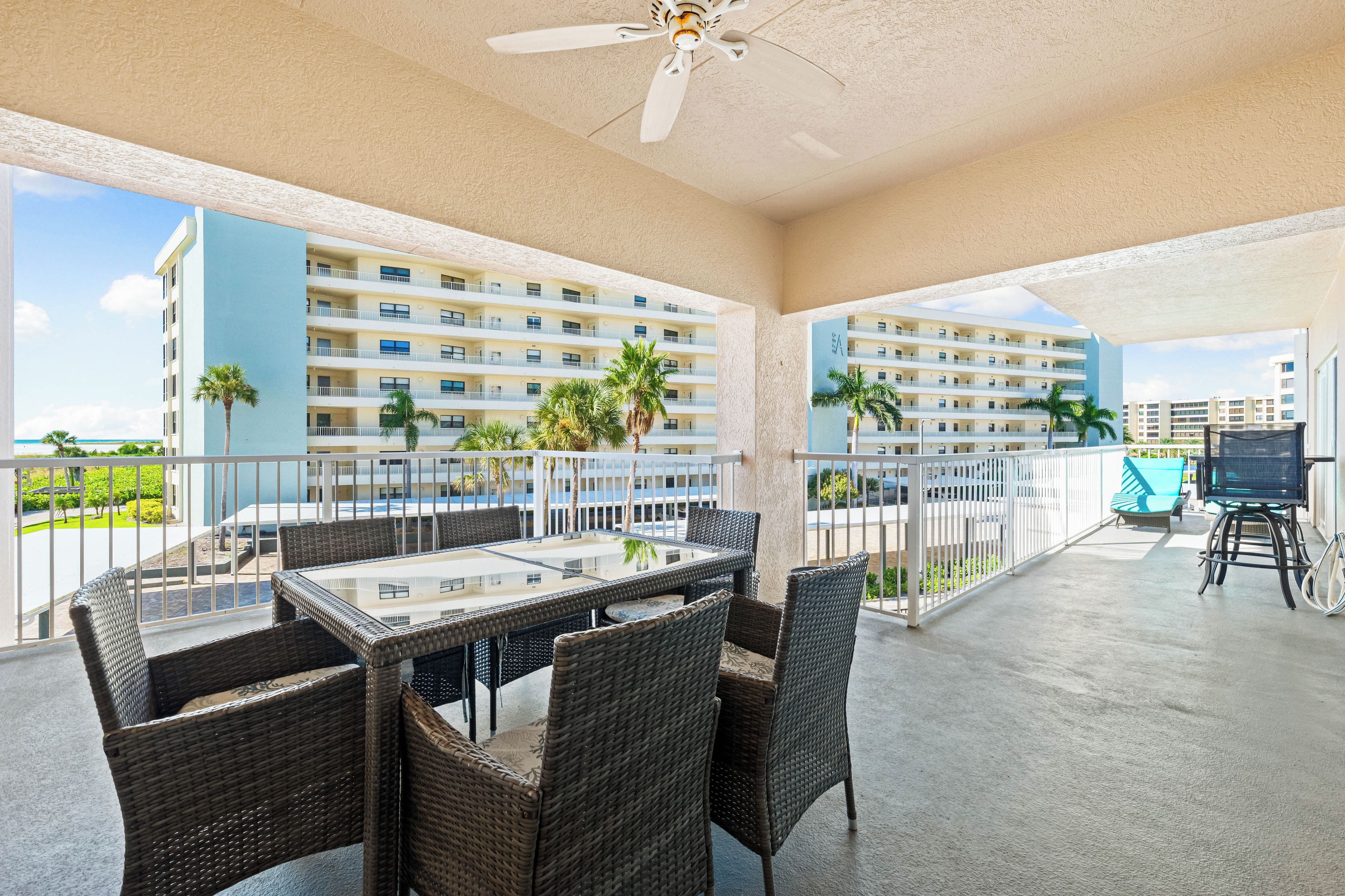 A balcony with wicker furniture, a ceiling fan, and a view of a multi-story building with balconies and palm trees.