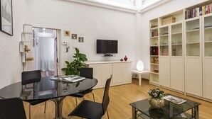 Uma sala de estar moderna com uma mesa redonda de vidro, cadeiras pretas, uma TV de tela plana, uma estante e um armário branco.