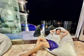A person reclining on a bean bag chair in a room with a glass wall, a pool, and a city view. - Best World Vision (Koh Samui)