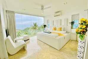 A modern bedroom with a large bed, a white sofa, a curved chair, a balcony with a view, and a vase with flowers.