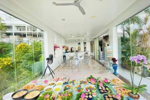 Un espacio interior amplio con una mesa larga que exhibe diversos alimentos, un ventilador de techo y vistas al exterior a través de grandes ventanas.