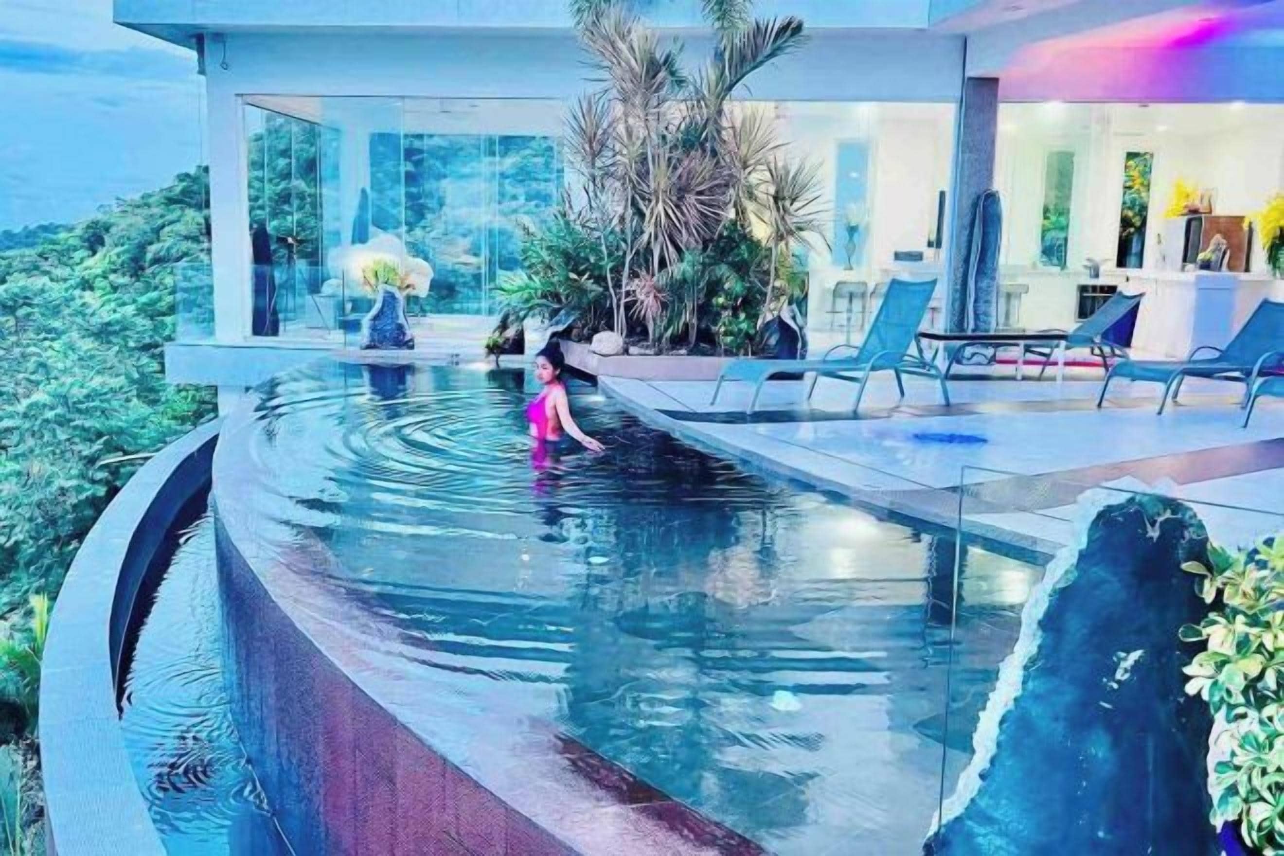 A pool with a waterfall feature, surrounded by lounge chairs and a glass-enclosed area with plants.