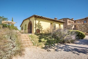 Ein Gebäude im mediterranen Stil mit einer Steintreppe, die zu einer Tür führt, umgeben von trockener Vegetation und blauem Himmel.