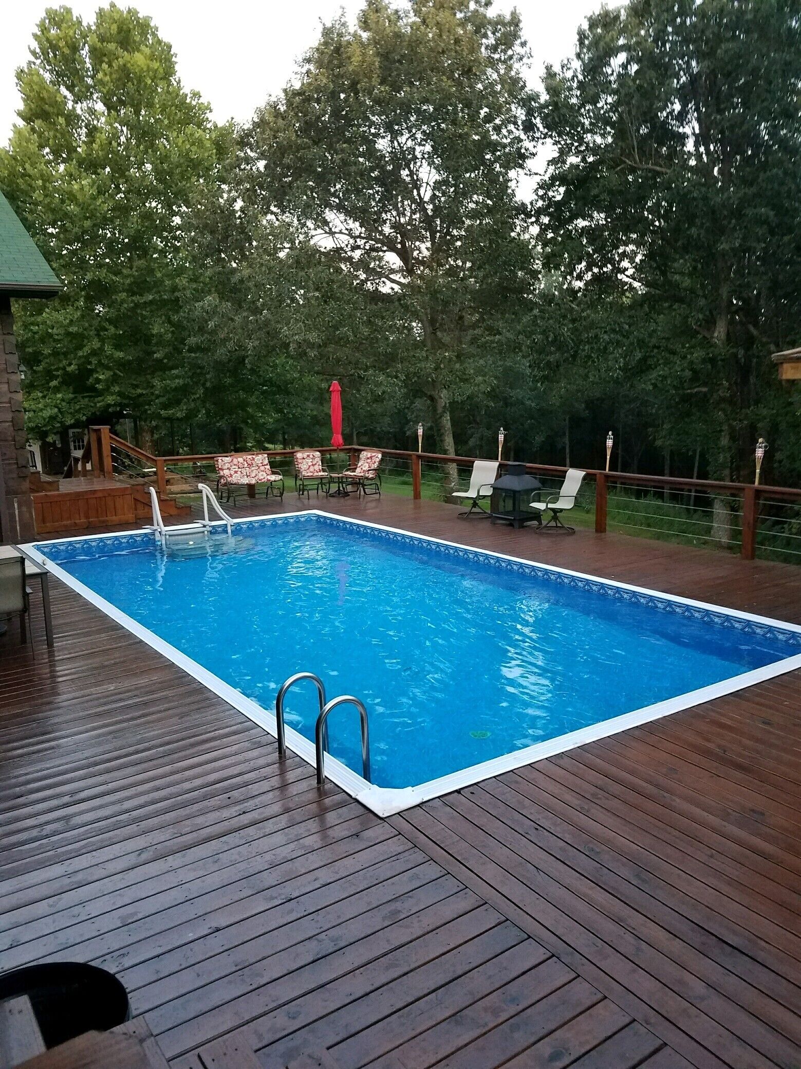 A rectangular swimming pool with a wooden deck, surrounded by trees and equipped with lounge chairs and a table.