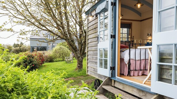 A cozy, single-room cabin with a wooden exterior, a small porch, and a view of a lush garden.