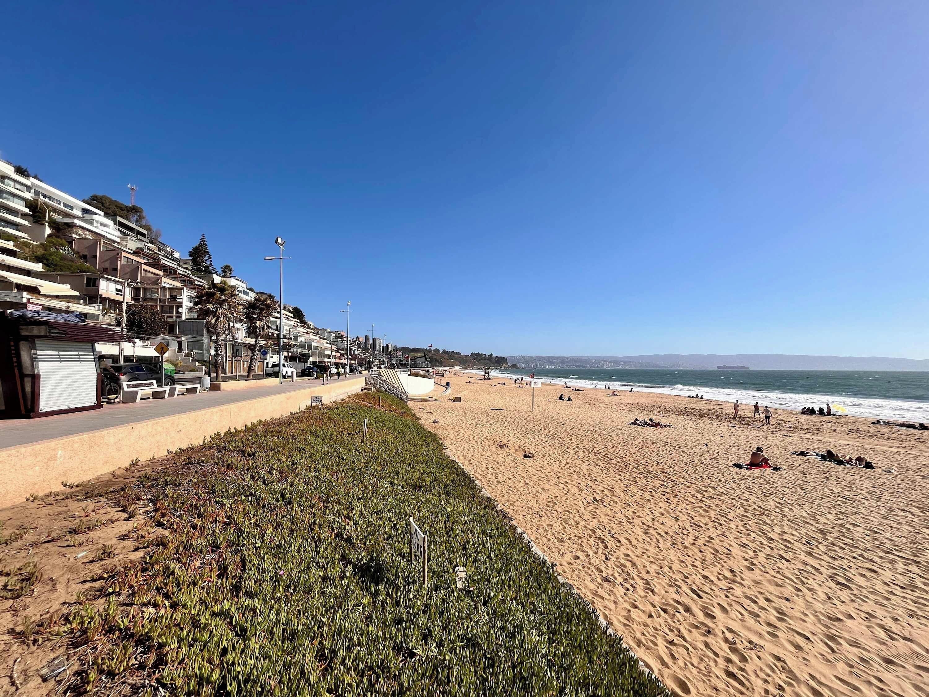 A coastal road with a beach on one side, lined with buildings and a clear sky.