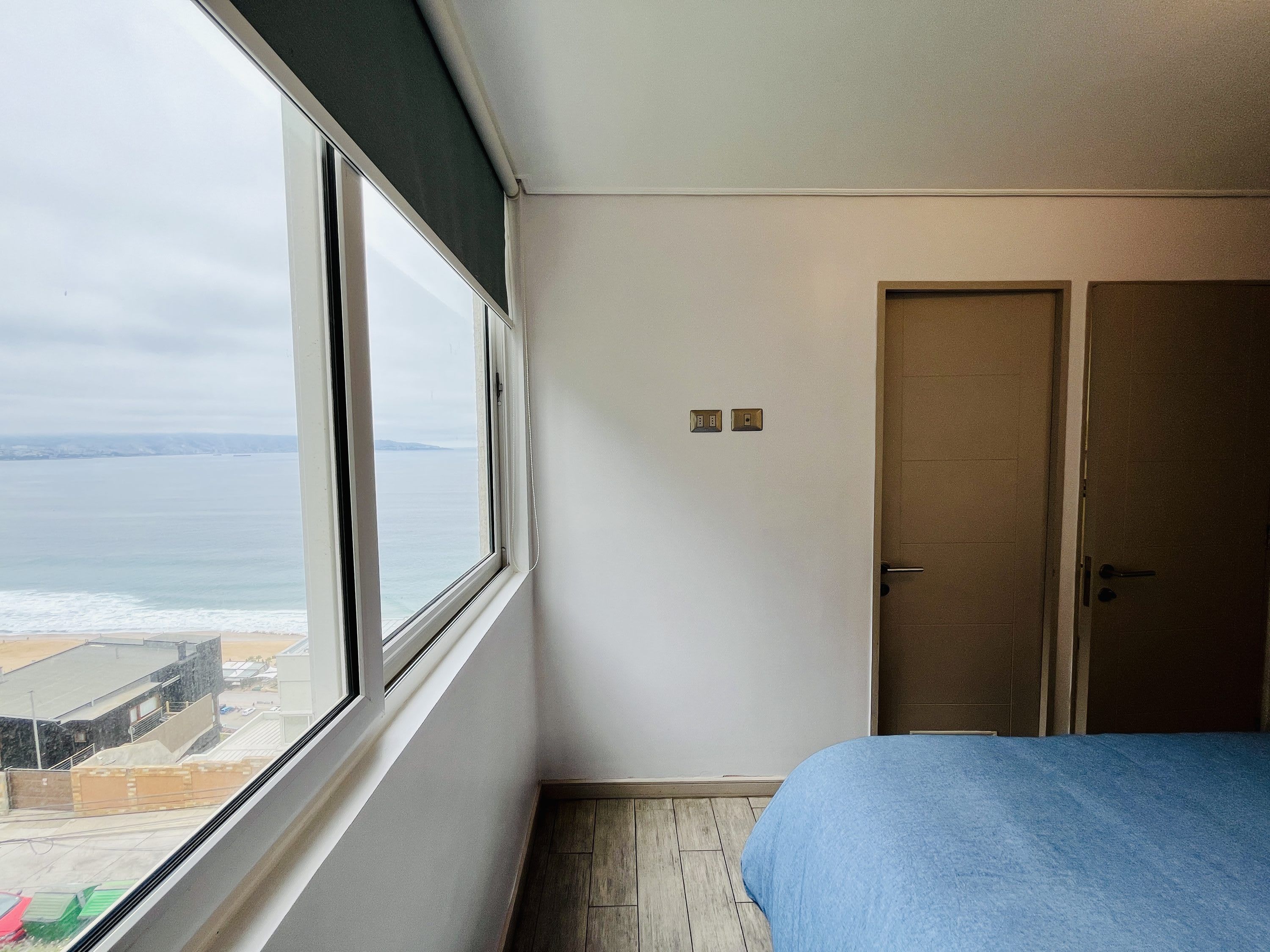 A room with a large window, a bed, a door, and a light switch on the wall.