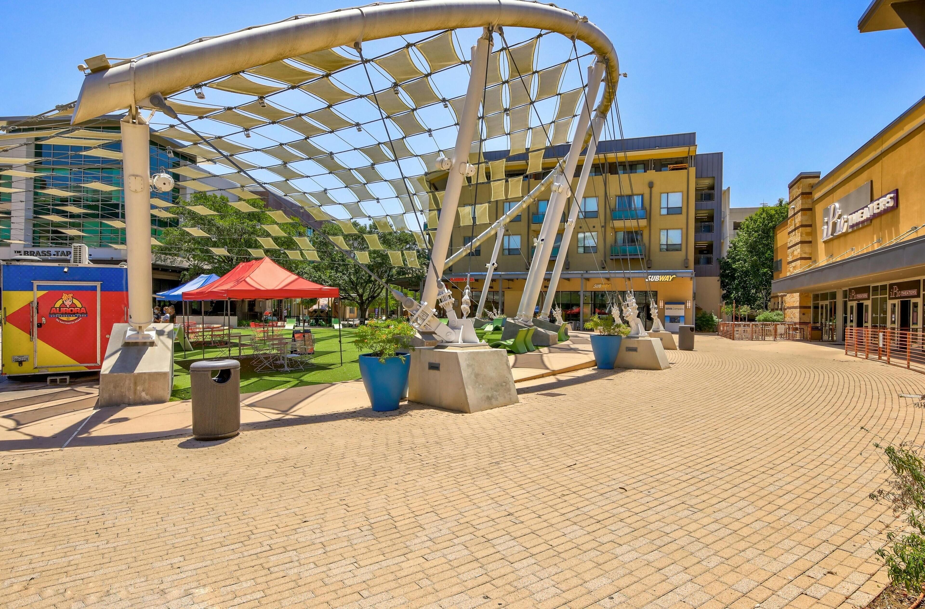 An outdoor plaza with a modern sculpture, seating areas, and a food court with visible restaurant signs.