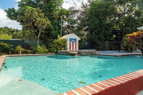 A swimming pool with a flagpole, surrounded by lush greenery and a stone wall. - Family House w. Pool, Parking & Basketball Court! In Rainforest!10 min to beach! (Rio Grande)