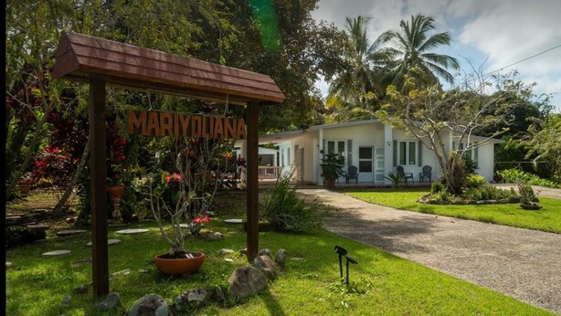 Una casa con un cartel que dice "MARIOLUANA", rodeada de exuberante follaje y palmeras.