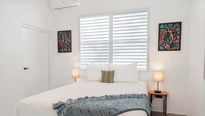 Un dormitorio con una cama, dos lámparas de noche, una ventana con persianas y dos obras de arte colgadas en la pared.