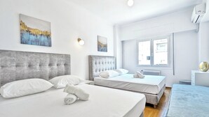 Habitación de hotel con dos camas, cabecero tapizado en gris, ropa de cama blanca, radiador y ventana con vistas.