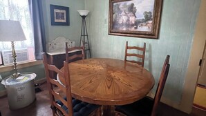 A dining area with a round wooden table and four chairs, a lamp, a picture frame, and a window with curtains. -  Sunday Haus- 2 blocks from Main Street! Sleeps 4 (Fredericksburg)