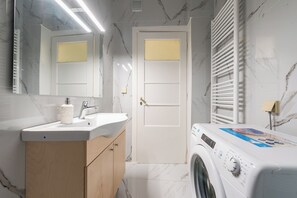 Uma casa de banho moderna com soalho de mármore, uma sanita branca, um armário de madeira, um espelho, uma porta e uma máquina de lavar roupa com abertura frontal.