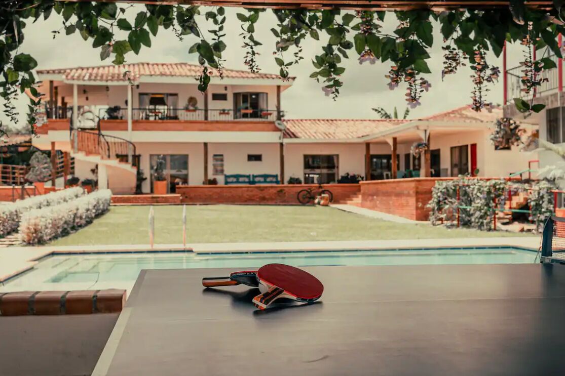 A pool table with a table tennis paddle and a cue ball, surrounded by a pool area with a view of a two-story building with balconies.