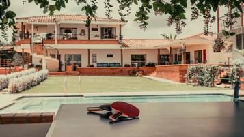 A pool table with a table tennis paddle and a cue ball, surrounded by a pool area with a view of a two-story building with balconies.