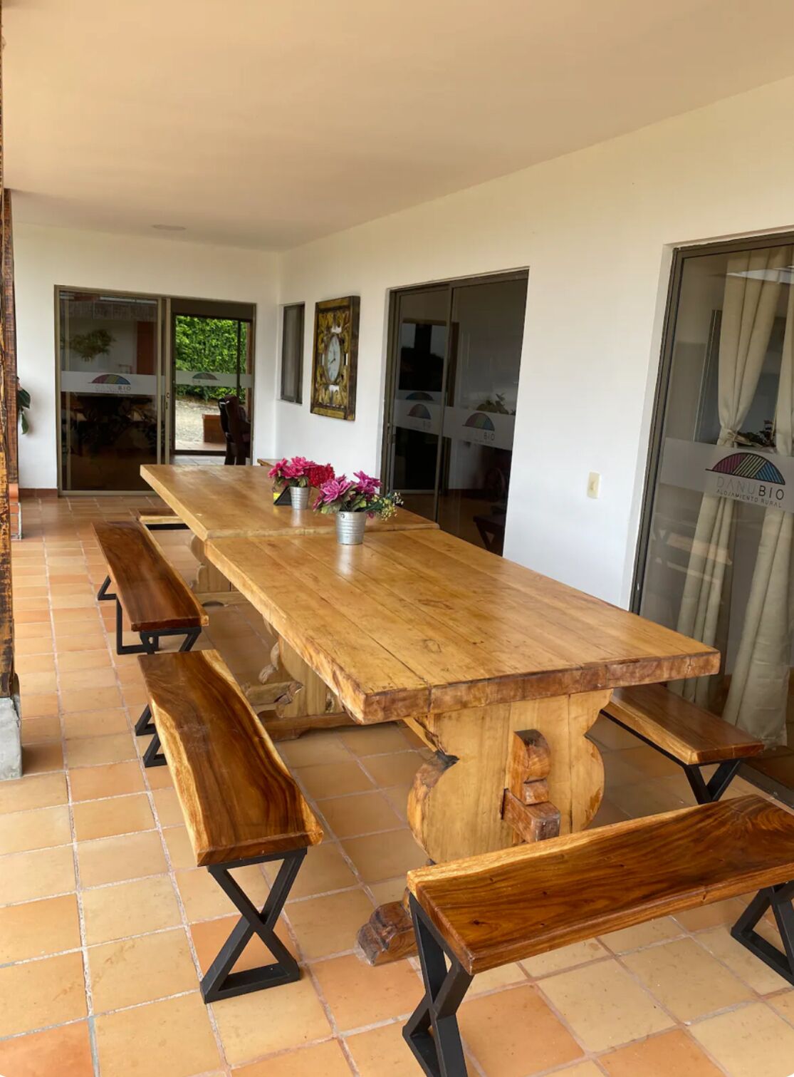 A long wooden dining table with benches, set on a tiled floor, in a room with white walls and a glass door leading outside.
