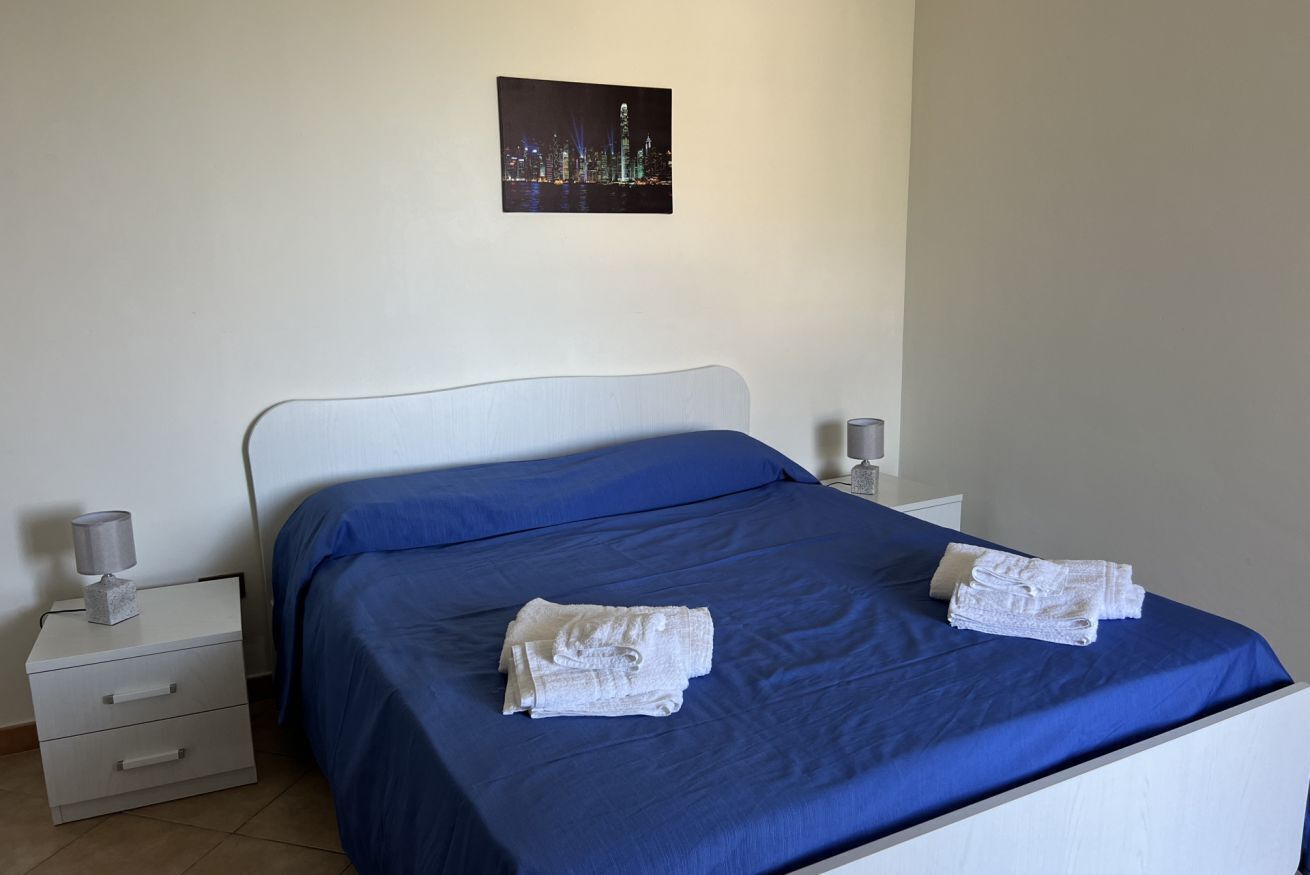 A neatly made bed with a blue bedspread and two folded white towels. There is a white nightstand with a lamp and a small decorative item on it. A framed picture of a cityscape is hung on the wall above the bed.