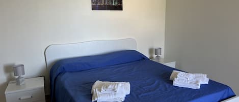 A neatly made bed with a blue bedspread and two folded white towels. There is a white nightstand with a lamp and a small decorative item on it. A framed picture of a cityscape is hung on the wall above the bed.