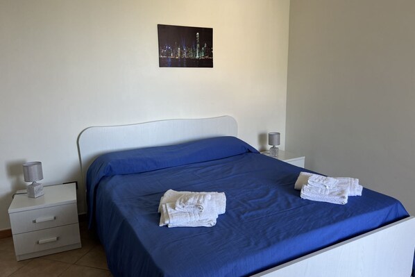 Una cama bien hecha con una colcha azul y dos toallas blancas dobladas. Se observa un mesita de noche blanca con una lámpara y un pequeño adorno. Encima de la cama, hay un cuadro enmarcado con una vista de la ciudad.
