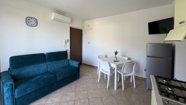 Un espace de vie compact comprenant un canapé de couleur turquoise, une petite table à manger avec des chaises, un téléviseur blanc fixé au mur et un coin cuisine avec un réfrigérateur et une cuisinière.