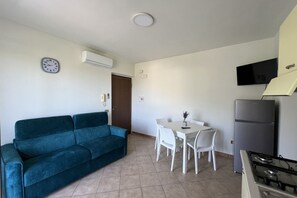 Un espacio habitable compacto con un sofá color turquesa, una pequeña mesa de comedor con sillas, un televisor blanco montado en la pared y una zona de cocina con refrigerador y estufa.