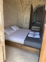 Una cama individual con sábanas blancas, una manta oscura estampada y dos toallas. La habitación tiene paredes de piedra y un marco de puerta de madera.