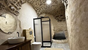 A rustic bathroom with a stone ceiling, a wooden vanity, a white sink, a toilet, and a black-framed shower enclosure. - Kochan Konagi (Mardin)