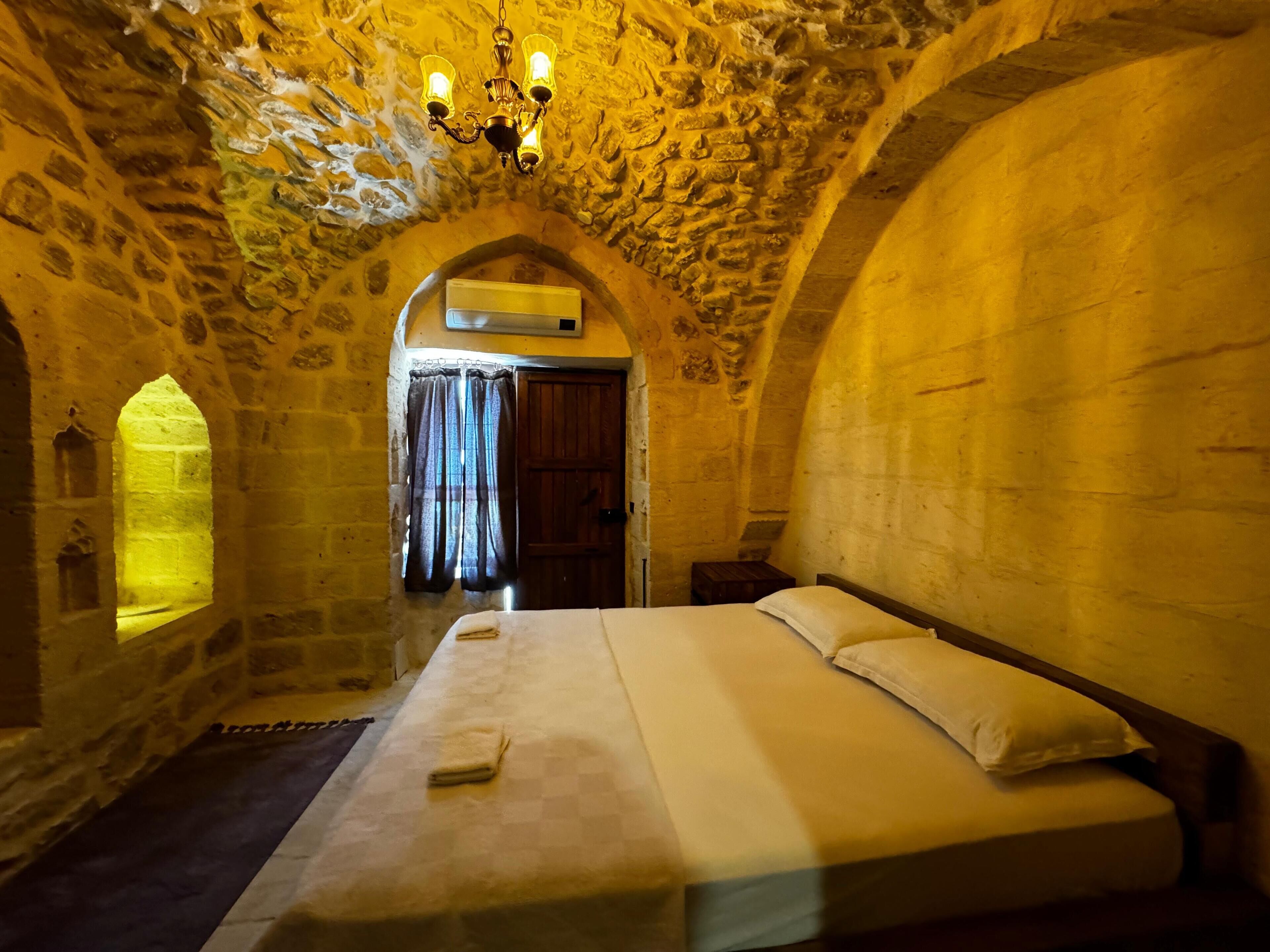 A stone-walled room with a bed, a chandelier, and an arched doorway.